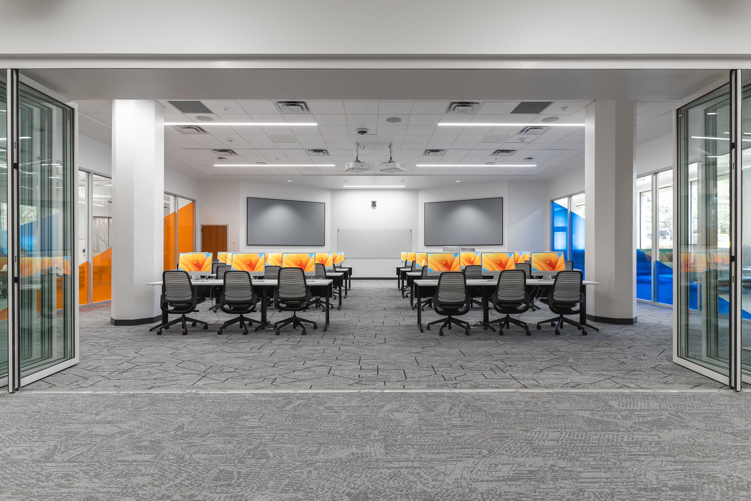 The image is of the Scholar's Lab. It shows eight tables with three computers at each table. The tables are facing the front wall of the lab, where there are two large screens.