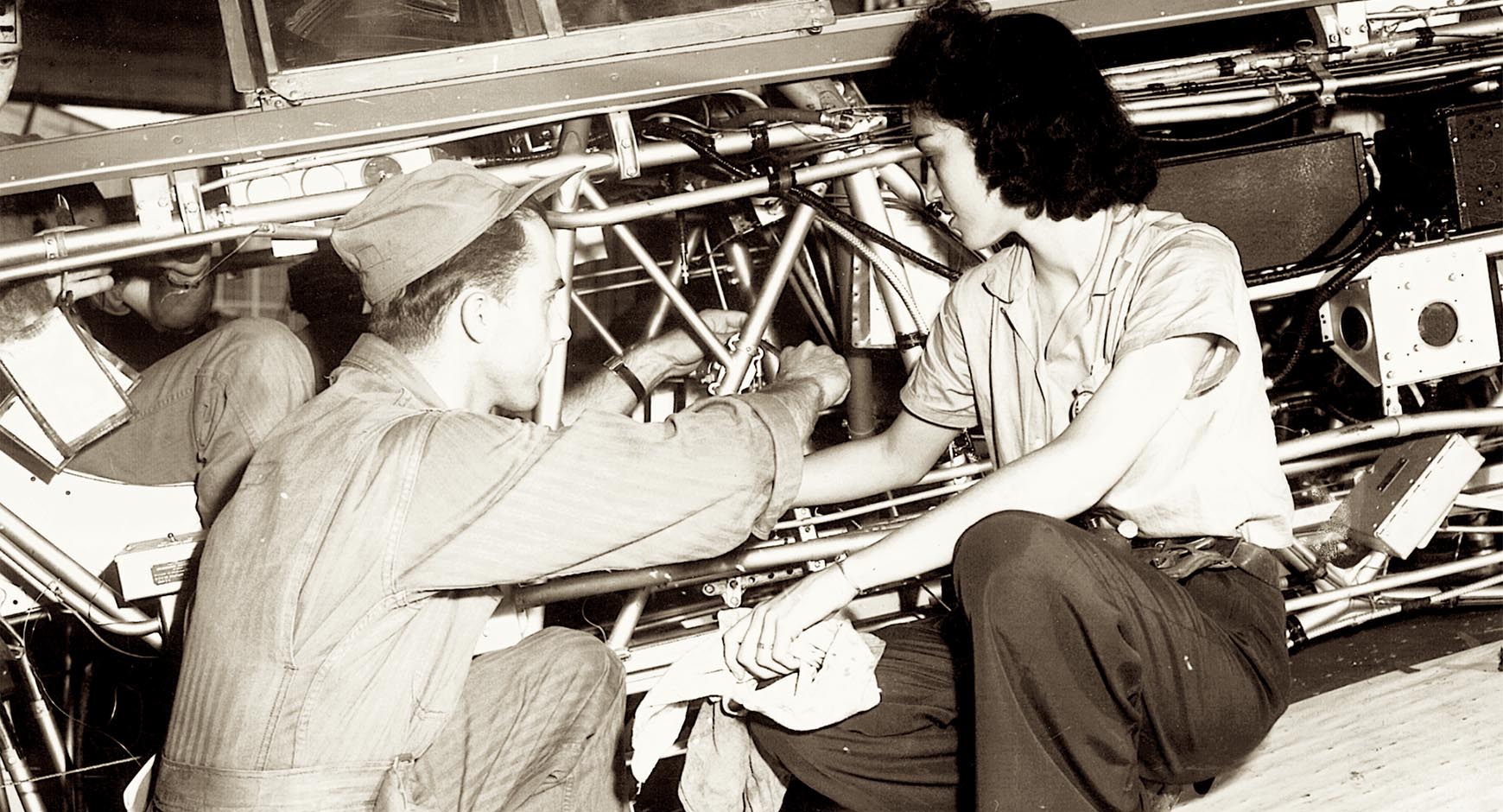 Black and white image of Latinx man and woman working on an assembly line, kneeling before an unidentifiable machine, both in work clothes.