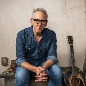 Man in glasses wearing denim sitting on bench next to guitar, hands clasped in front of him.